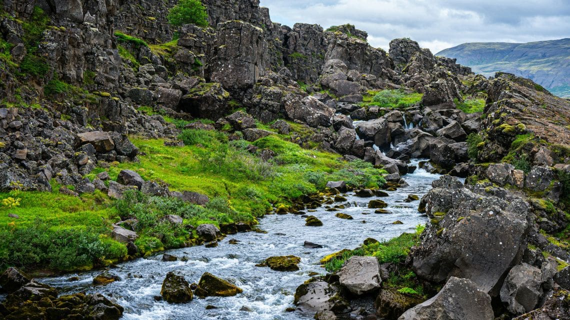 A small, clear stream flows through a rocky landscape surrounded by vibrant green vegetation. Rugged cliffs rise in the background under an overcast sky.
