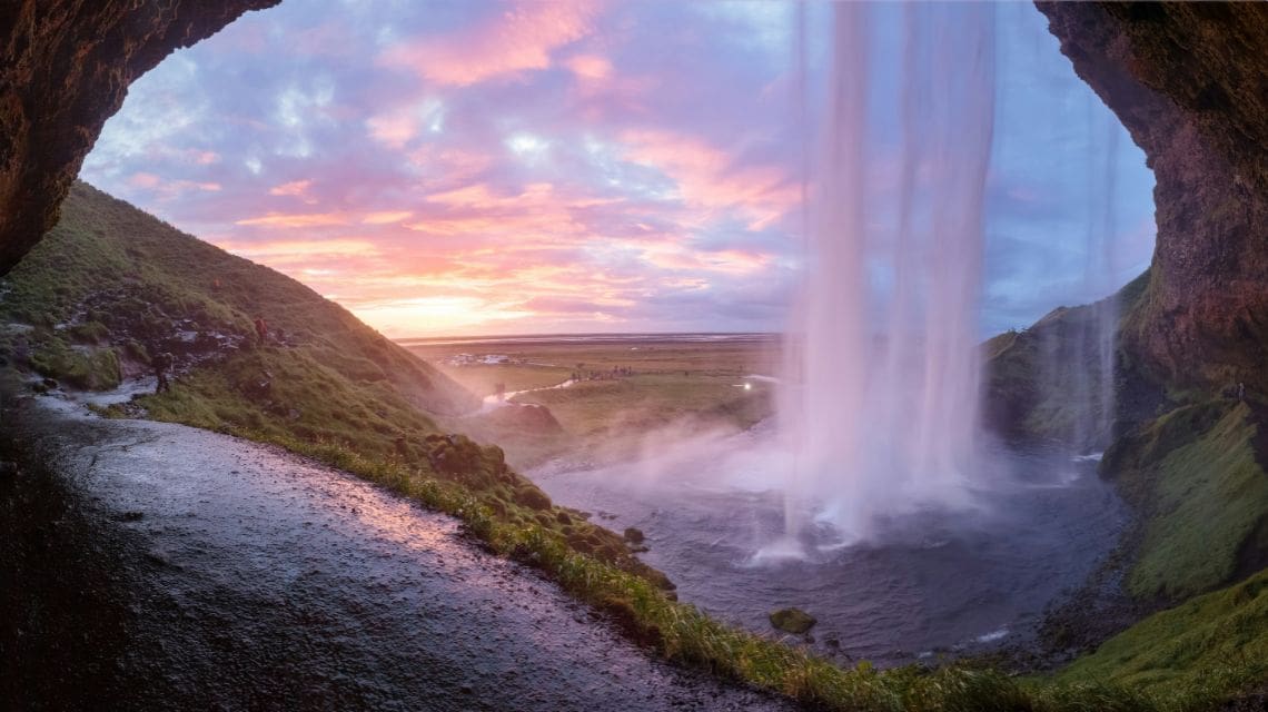 View from behind a waterfall, with a sunset sky painting vibrant pink and orange hues over a tranquil landscape. Water cascades gently, creating a serene, dreamy scene.