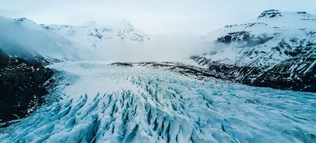 A vast, rugged glacier with deep crevices stretches across a mountainous landscape. Snowy peaks and misty clouds create a serene, cold atmosphere.