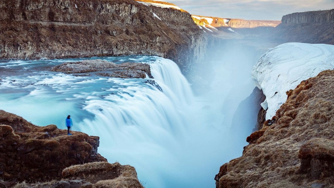 A person in a blue jacket stands on a cliff edge, gazing at a majestic waterfall cascading into a misty gorge. Snow and rocky terrain surround the scene.