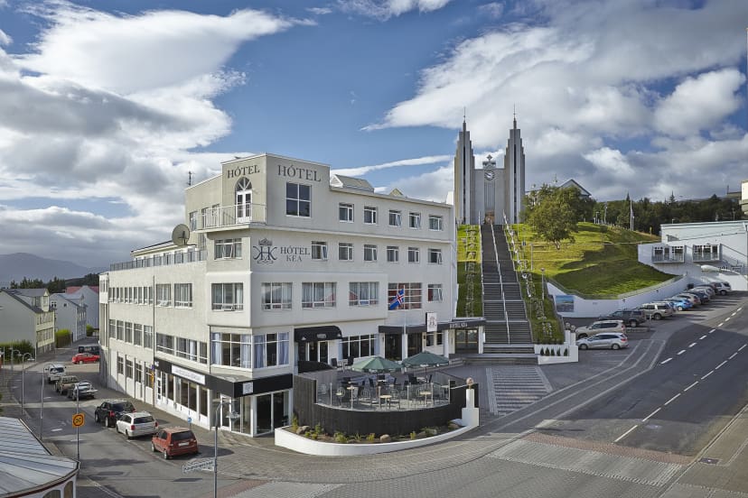 White hotel on a corner under a blue sky, with a terrace cafe outside. Behind it, steps lead up a hill to a modern church, surrounded by greenery.