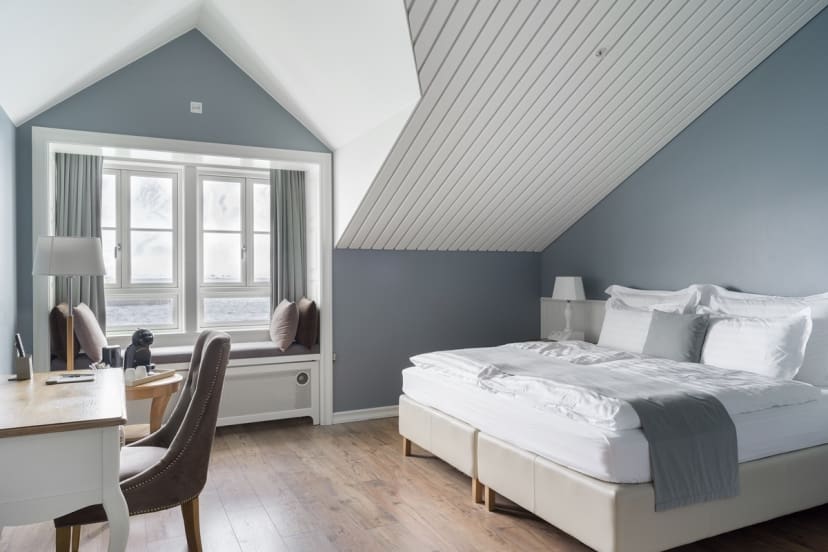 A serene bedroom with slanted white-paneled ceiling, blue-gray walls, and wood floor. Features a cozy bed, desk, chair, and a window with pillows.