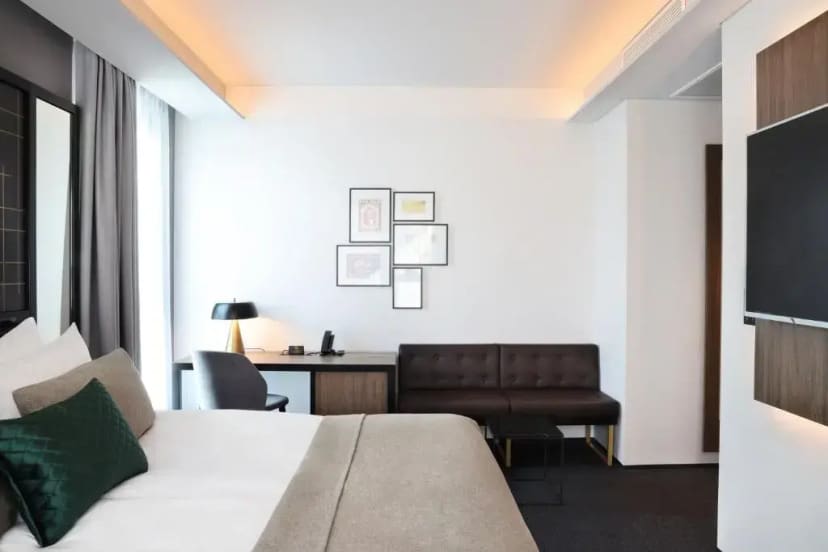 Modern hotel room with a large bed featuring white and beige linens, a green cushion, a sleek desk, a leather sofa, and framed art on a white wall.