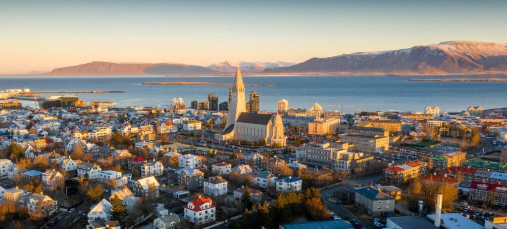 Aerial view of Reykjavik at sunset, featuring a prominent cathedral with a tall spire, surrounded by colorful houses. Mountains and water in the background.