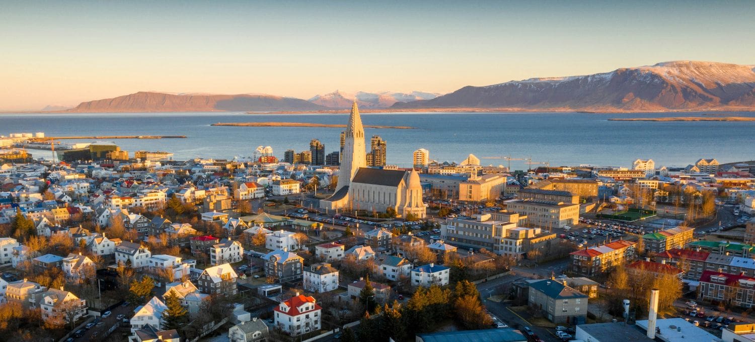 Aerial view of Reykjavik at sunset, featuring a prominent cathedral with a tall spire, surrounded by colorful houses. Mountains and water in the background.