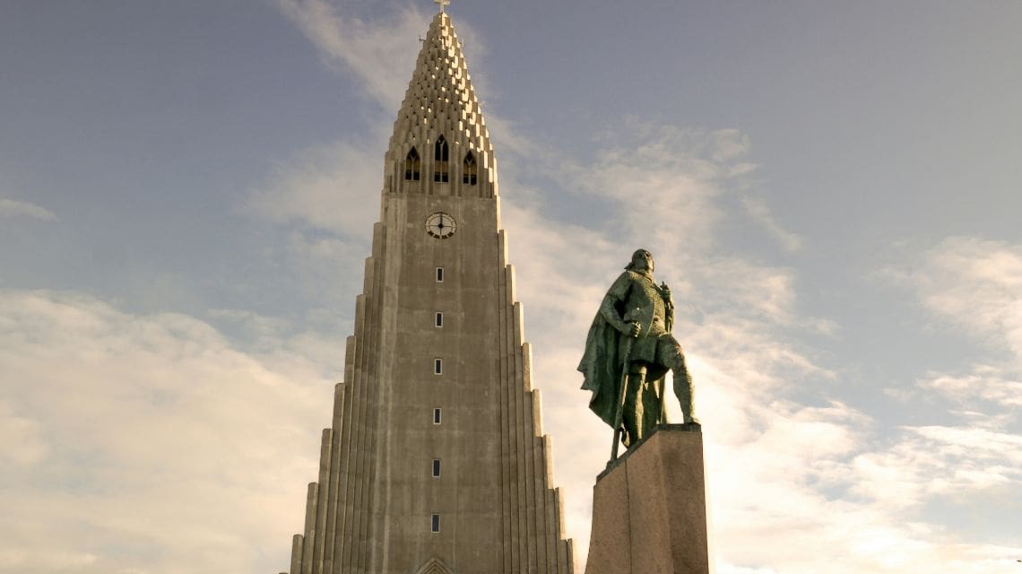 A tall, angular church tower rises under a cloudy sky. A statue of a figure in a cape stands on a pedestal in the foreground, conveying grandeur.