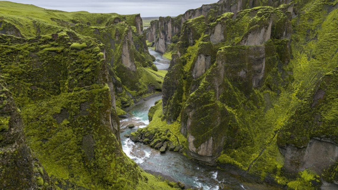 Aerial view of a lush, green canyon with steep cliffs covered in vibrant moss. A winding river flows through the canyon, creating a serene and majestic landscape.