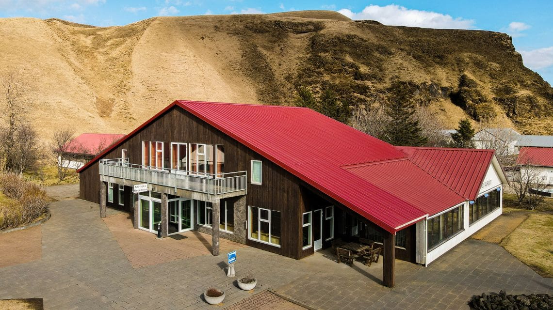A rustic building with a vibrant red roof and wooden siding sits in front of a rugged hillside under a blue sky. The setting is serene and remote.