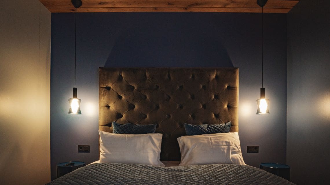 Cozy bedroom with dim lighting, features a tufted headboard, two pillows, and a quilted bedspread. Warm pendant lights hang on both sides.