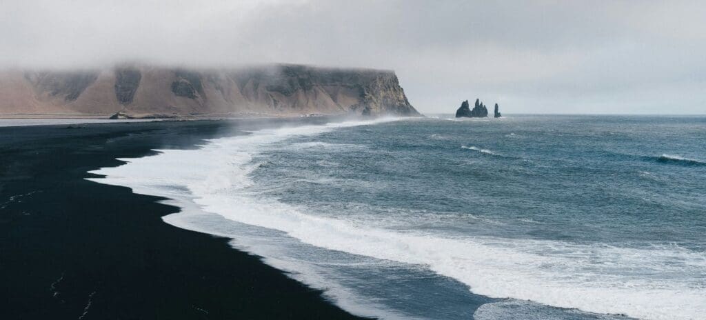 A misty view of a black sand beach with gentle waves and distant sea stacks. The rocky cliffs are shrouded in fog, creating a serene and mystical atmosphere.