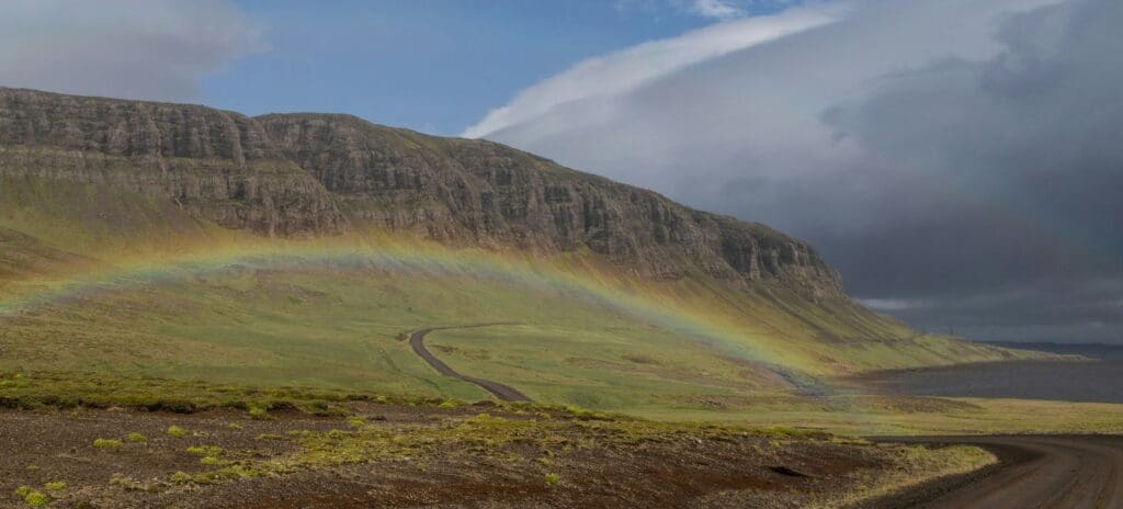 A curving dirt road runs through a vast, green landscape under a cloudy sky. A rainbow arches over the hills, adding a serene and hopeful touch.