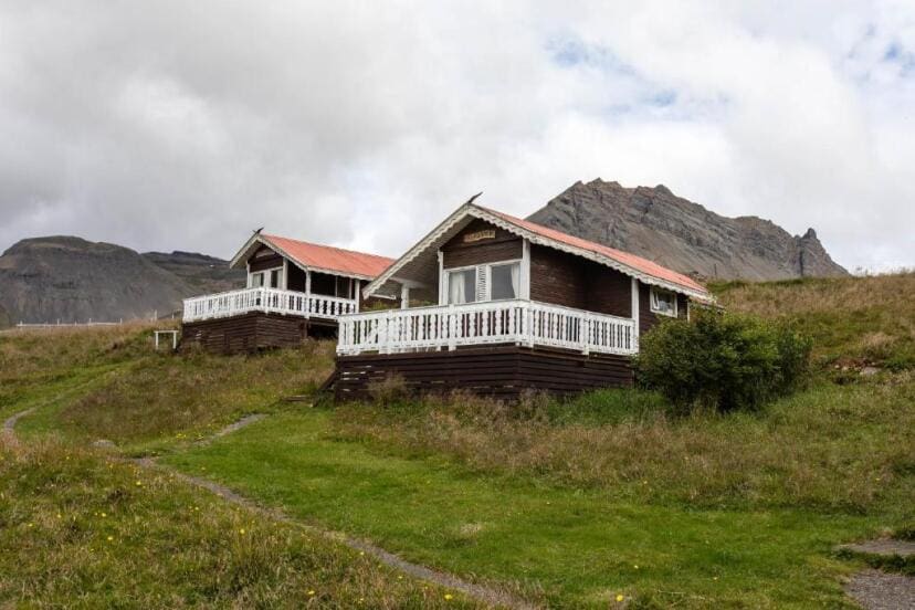 Two rustic wooden cabins with white trim sit on a grassy hillside, backed by rugged mountains under a cloudy sky, creating a serene, remote atmosphere.