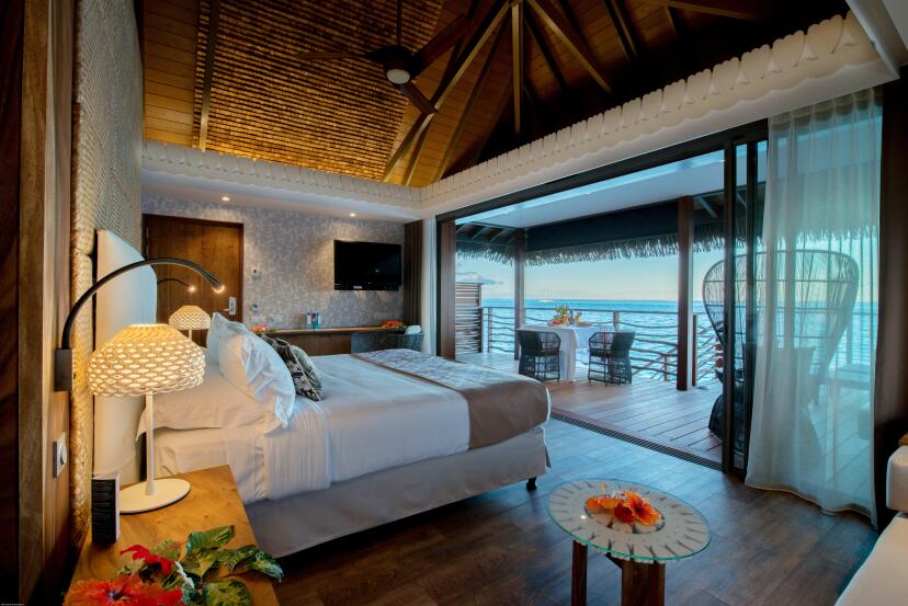 Luxurious tropical hotel room with a wooden ceiling, a cozy bed, and a view of the ocean. An open sliding door leads to a patio with seating.