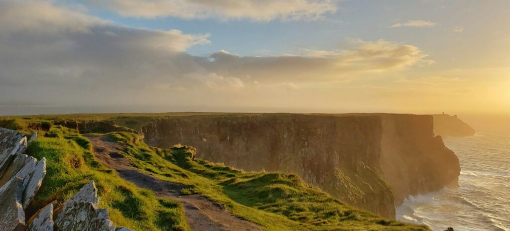 Cliffs at sunset with a rocky path and lush green grass, overlooking the ocean. Sunlight bathes the scene warmly, creating a serene and majestic atmosphere.