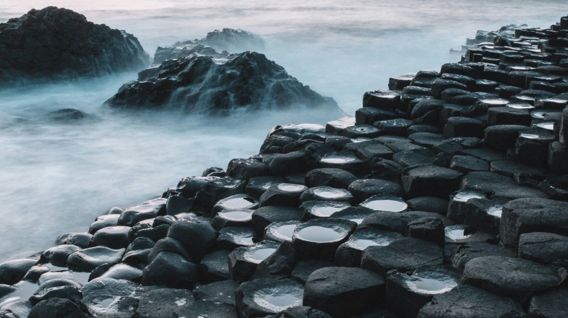 Rocky coastal scene with hexagonal basalt columns, wet from ocean spray, leading into misty waves at dawn. Moody, serene atmosphere.