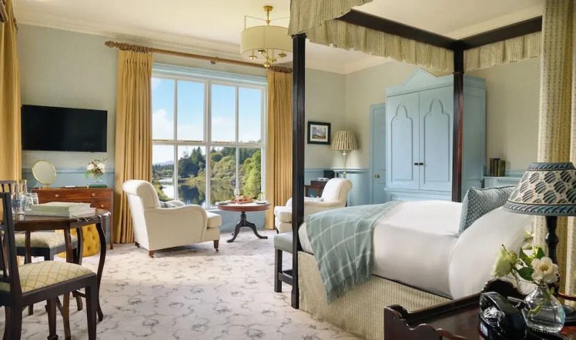 Luxurious bedroom with a four-poster bed, light blue and cream decor, elegant seating by large window offering a scenic view of trees under a blue sky.