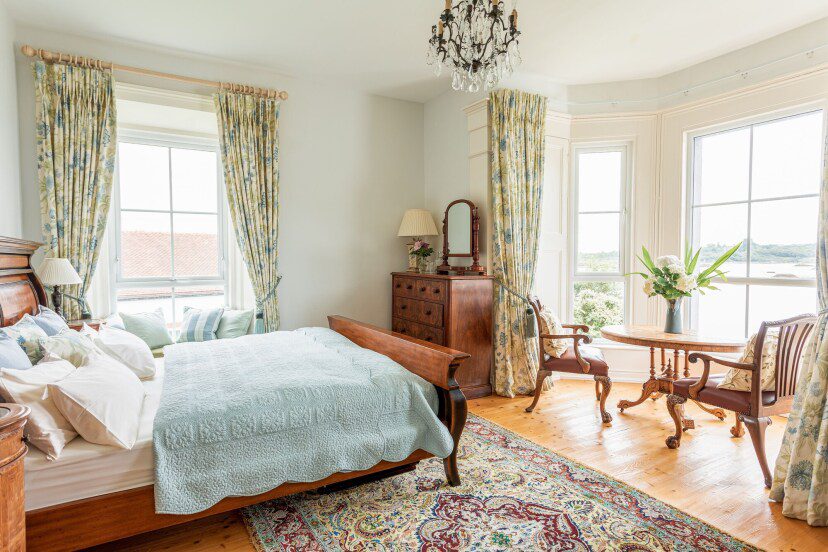 Elegant bedroom with large windows and floral curtains, featuring a wooden bed with a blue cover, antique furniture, chandelier, and scenic views.