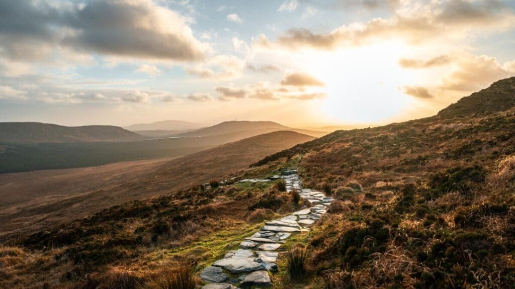 A winding stone path leads through a rugged landscape under a golden sunset, with hills and sparse vegetation creating a serene and expansive view.
