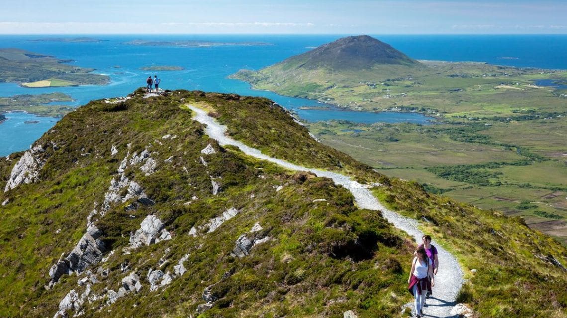 A winding trail on a lush green mountain leads to hikers, overlooking a vast blue ocean and distant islands. The scene is serene and expansive.