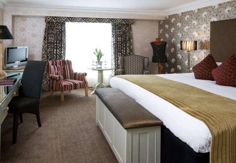 Stylish hotel room with a large bed, gold throw, and red pillows. Elegant wallpaper, striped and patterned chairs, a desk with a computer, and a mannequin create a cozy, sophisticated atmosphere.