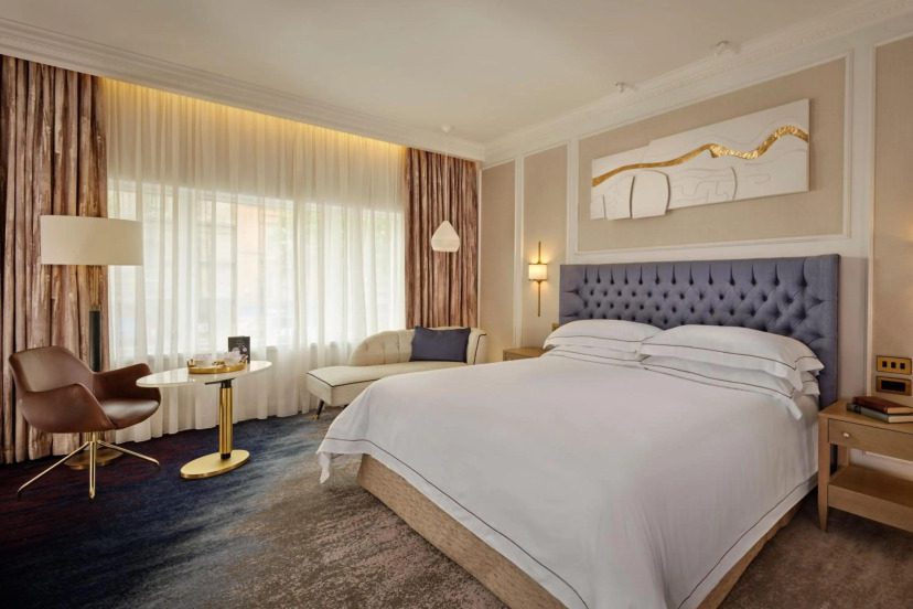 Elegant hotel room with a king bed, tufted headboard, modern art, marble lamp, and seating area by large curtains. Soft lighting conveys luxury.