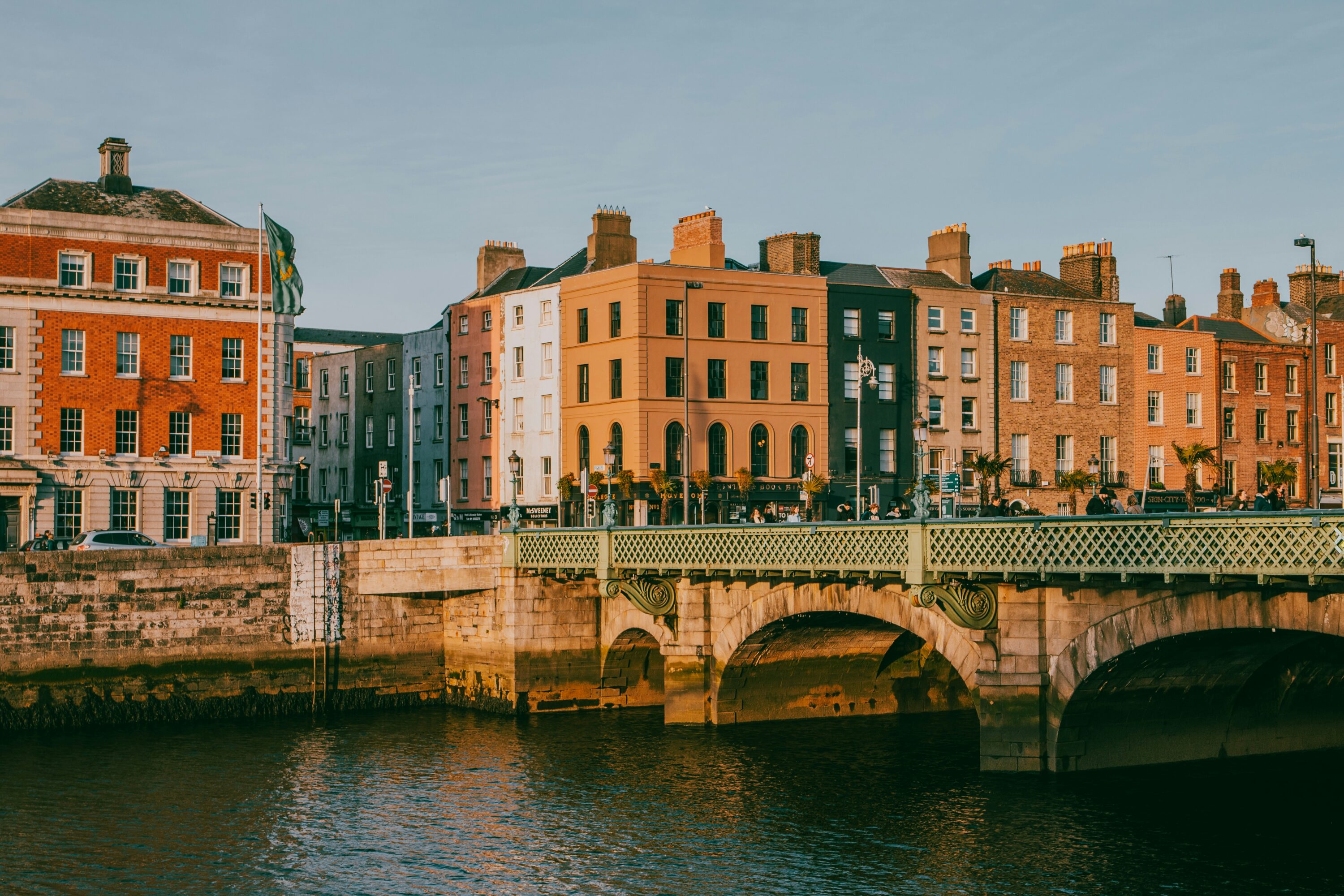 Historic European street along a river, featuring ornate bridge and colorful Georgian-style buildings lit by warm, golden sunlight. Calm and picturesque.