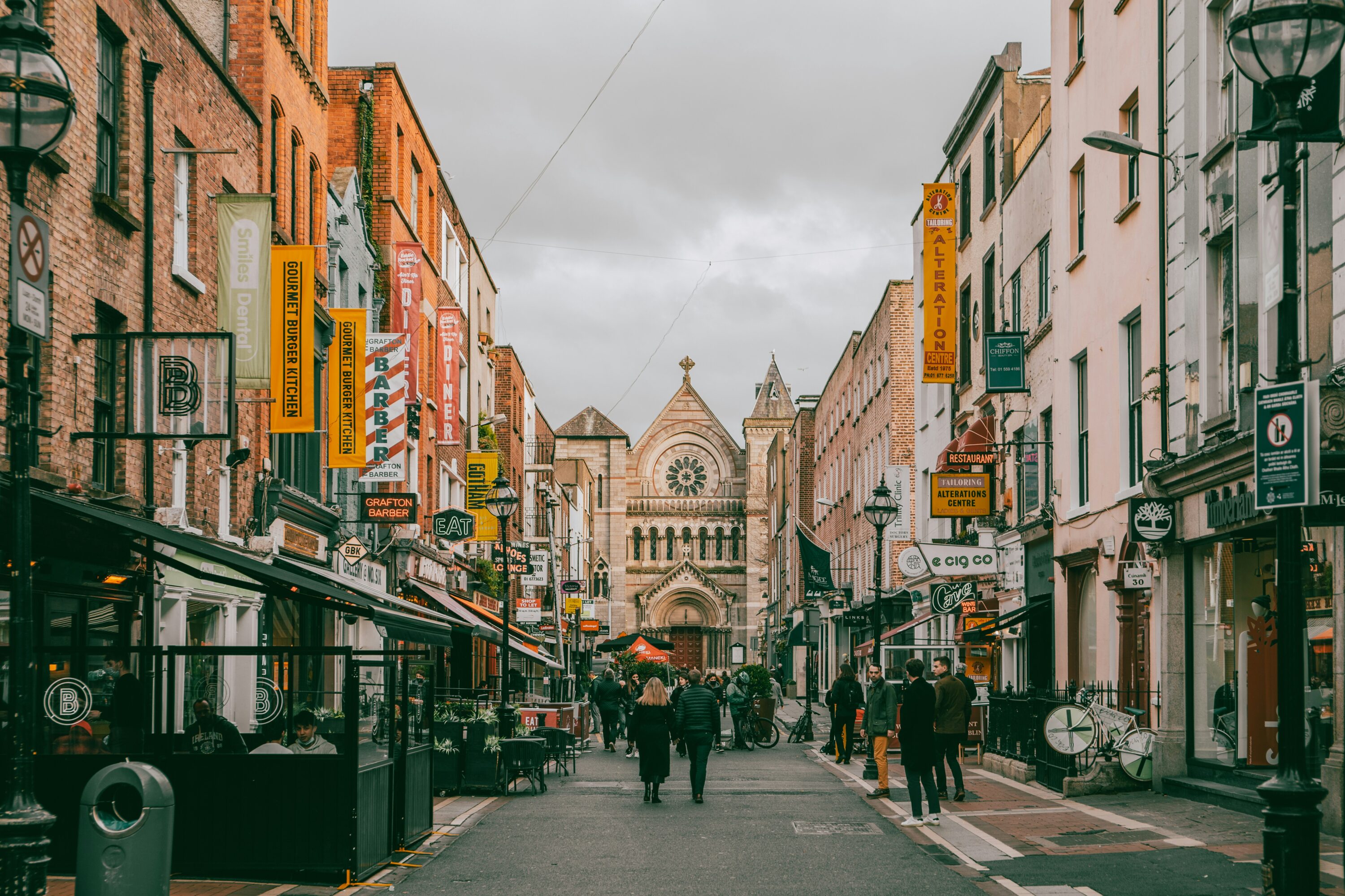 A bustling street scene with vibrant shops lining both sides, leading to a historical stone church under a cloudy sky. People stroll along the lively avenue.