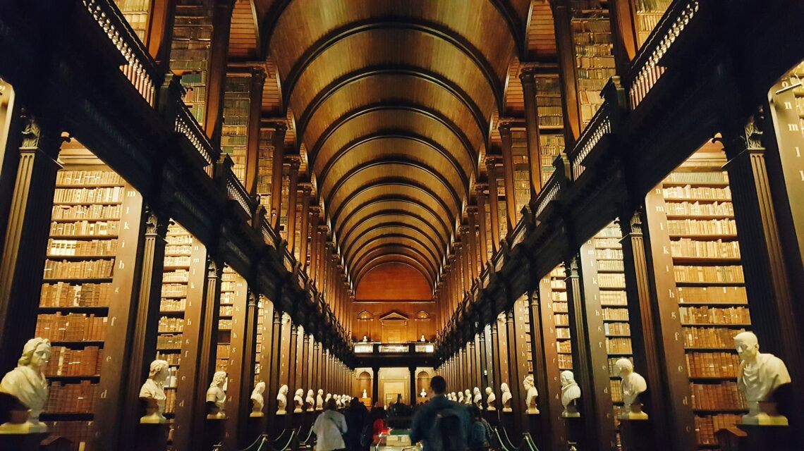 A grand library hall with a high, arched wooden ceiling, lined with tall shelves of books. White marble busts are displayed along the sides, and people are walking down the central aisle.