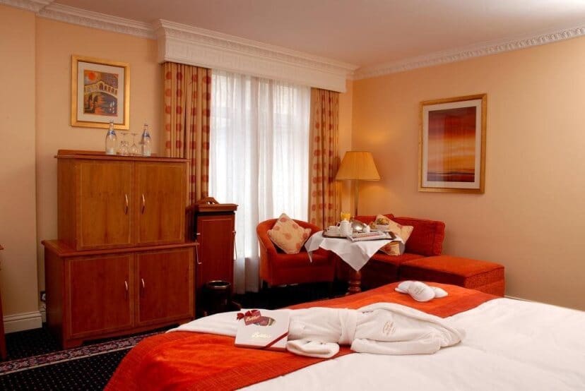 Cozy hotel room with a large bed draped in white and orange linen. Seating area with red chairs, table with breakfast, and soft lighting create a warm ambiance.