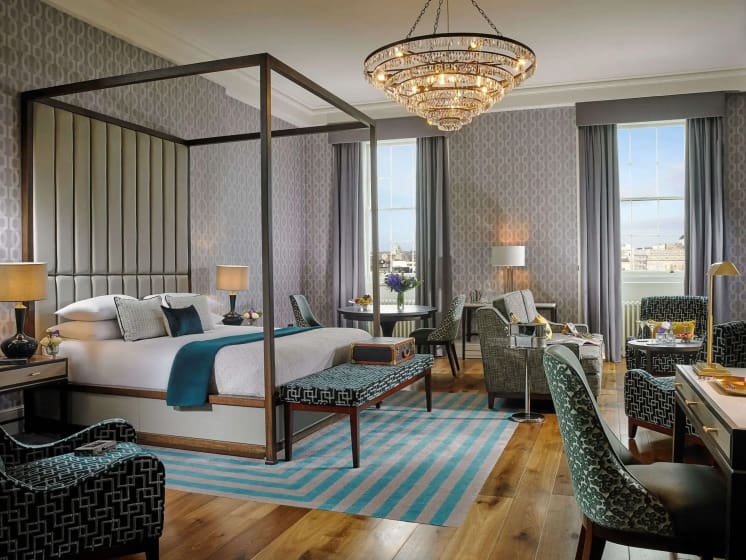 Elegant hotel room with a four-poster bed, blue accents, patterned chairs, and a large chandelier. Two tall windows reveal a city skyline.