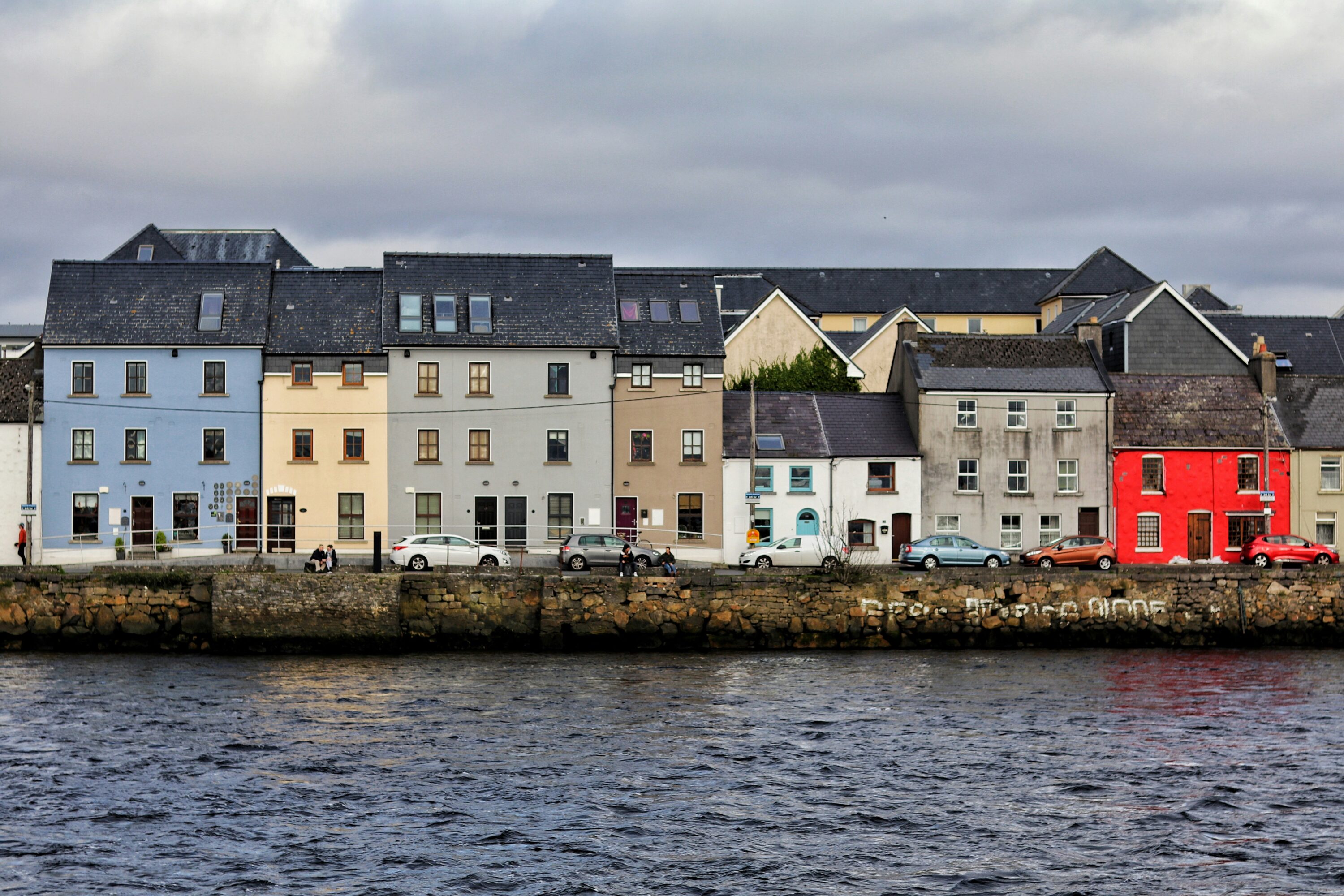 A row of colorful houses in varied shades of blue, yellow, and red faces a river under a cloudy sky. The scene conveys a quaint, serene coastal charm.
