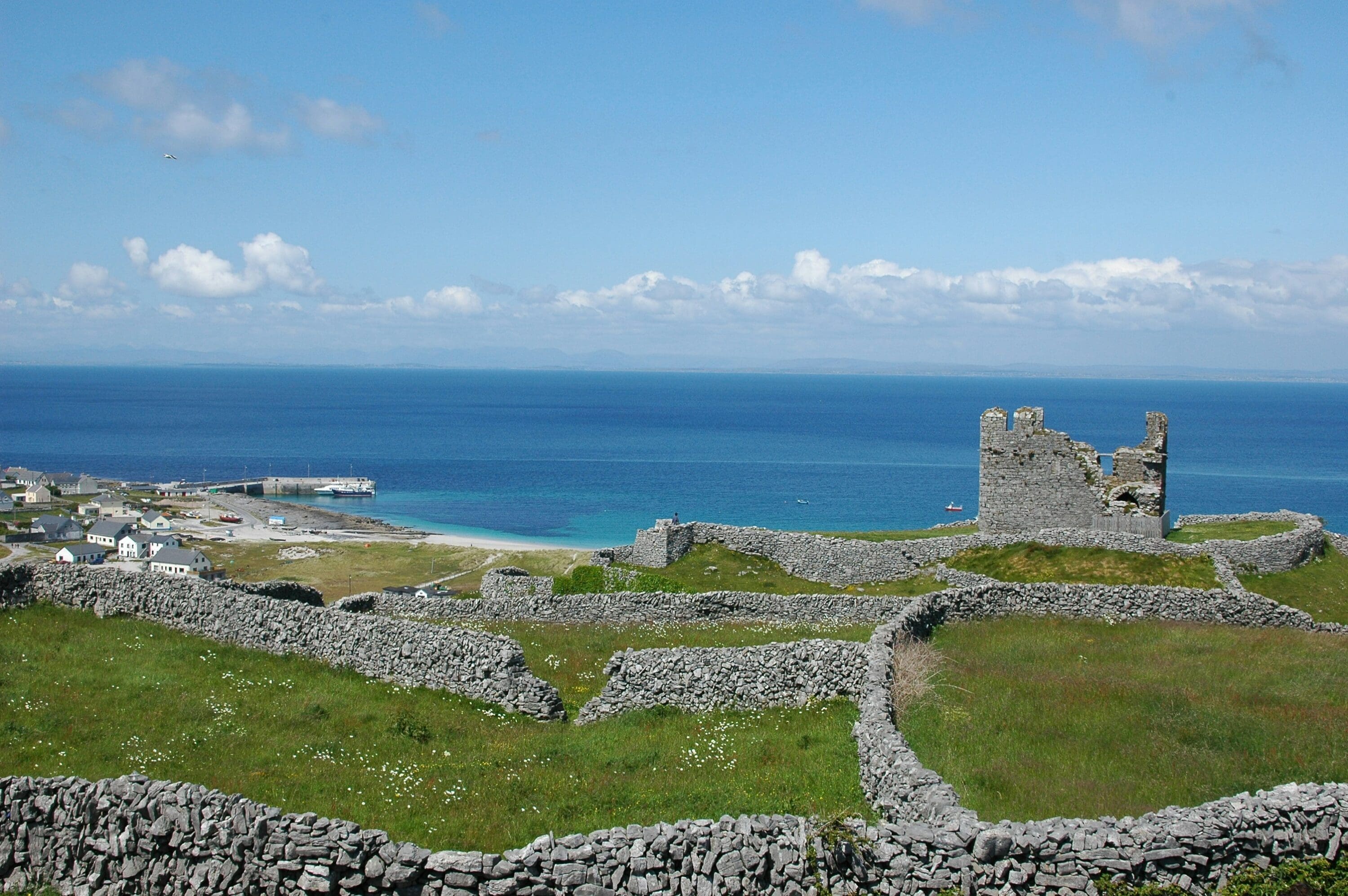 Scenic coastal landscape with ancient stone ruins on grassy hills, overlooking blue ocean and distant horizon. Clear sky with scattered clouds. Peaceful ambiance.