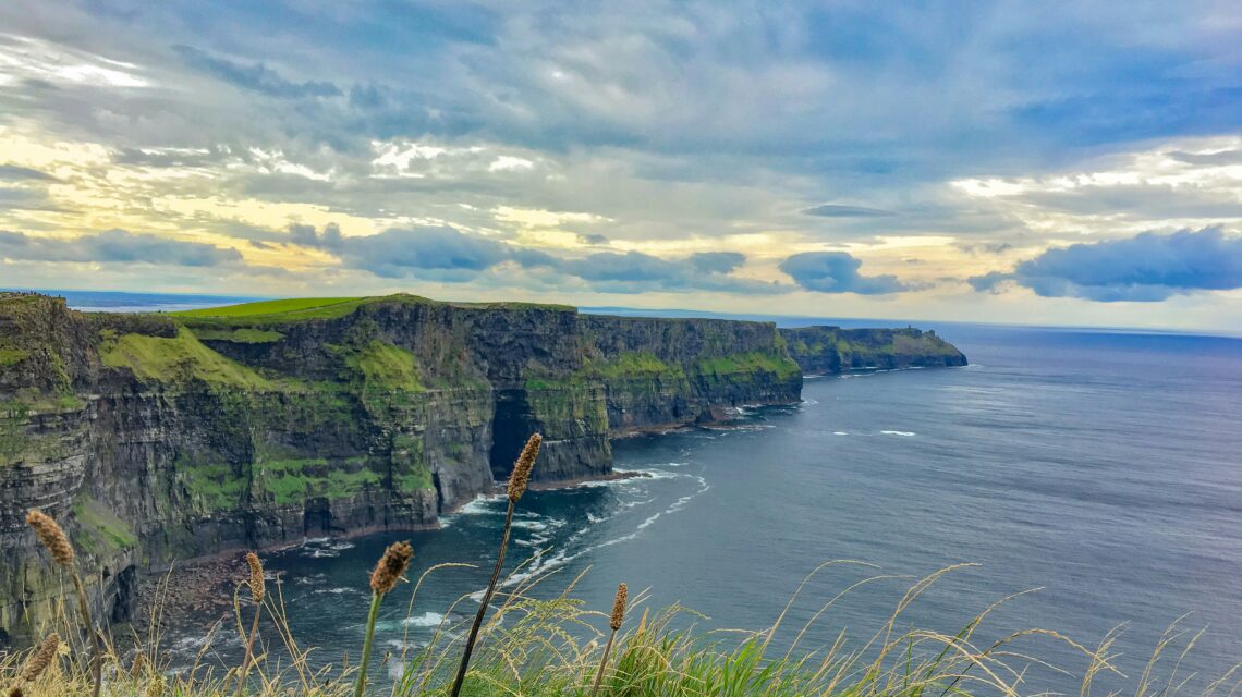 Majestic cliffs with lush greenery overlook a vast ocean under a dramatic sky. Foreground contains tall grasses, adding depth. Serene and awe-inspiring.