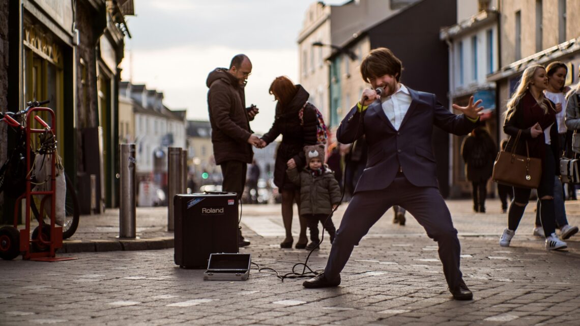 Street performer passionately sings in a lively pedestrian area, wearing a suit. Bystanders, including a family and a child, watch and stroll by.