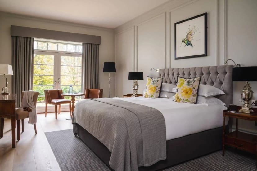 Elegant hotel room with a king-sized bed, gray bedding, and floral pillows. Sunlight filters through large windows, creating a warm, inviting ambiance.