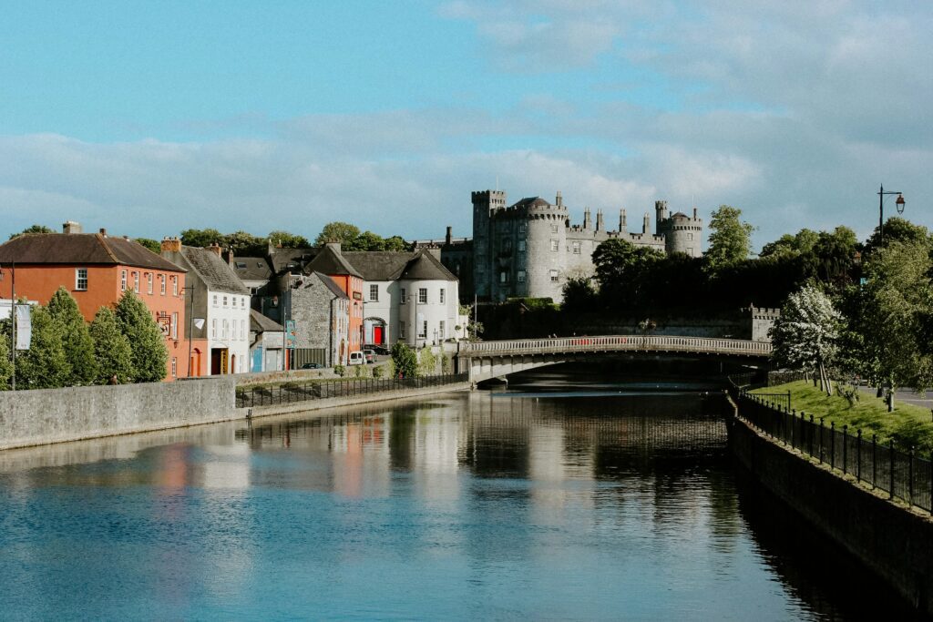 A serene riverside scene with colorful historic buildings and lush greenery leading to a grand, medieval-style castle under a bright blue sky.