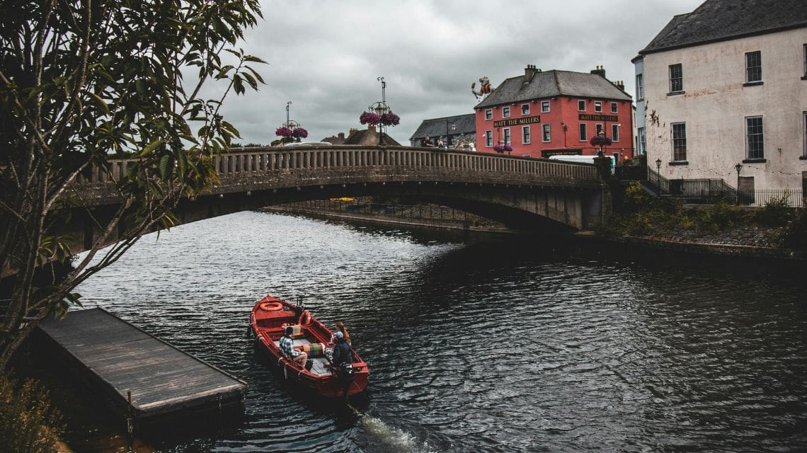 A red boat with two passengers floats on a calm river under a stone bridge. In the background, colorful buildings and overcast skies add a peaceful tone.