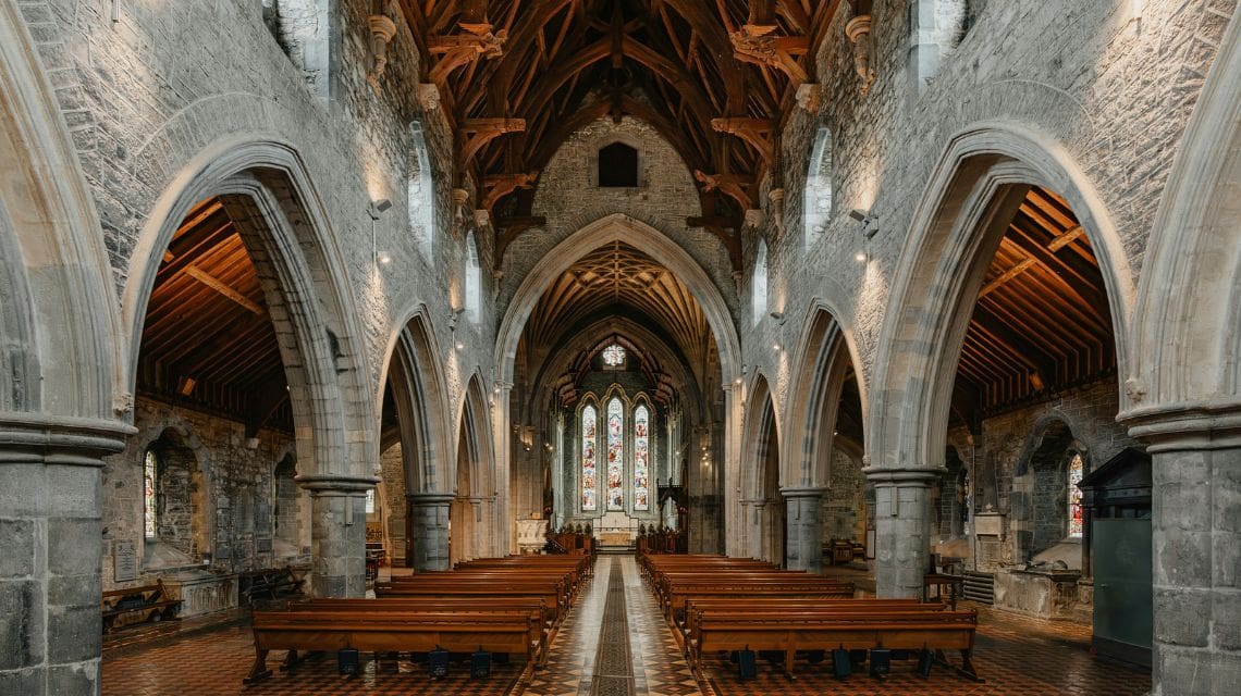 Majestic church interior featuring stone arches, wooden pews, and a high vaulted ceiling. A stained glass window at the back adds colorful light.