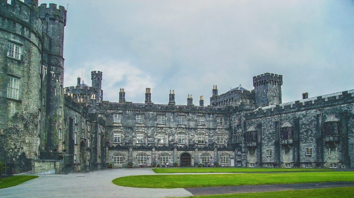 Gray stone castle with turrets, arched windows, and a green lawn under a cloudy sky. The scene conveys a historic and majestic atmosphere.