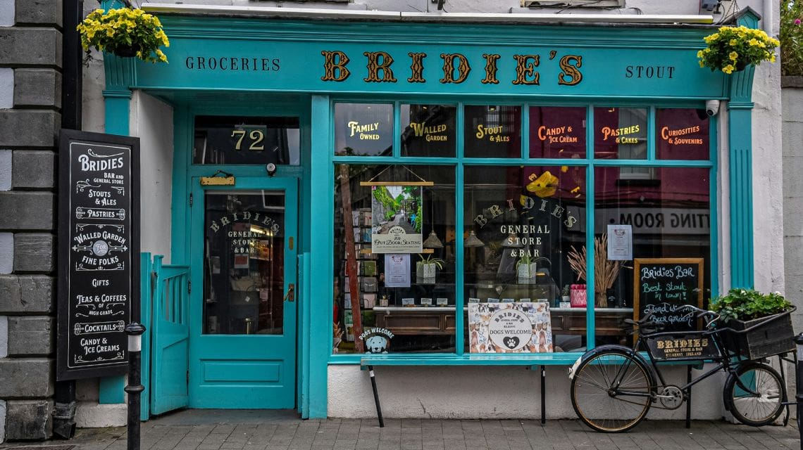 Charming storefront of Bridie's General Store and Bar with turquoise accents, vintage signage, potted flowers, and a bicycle with a woven basket outside. Warm, nostalgic ambiance.