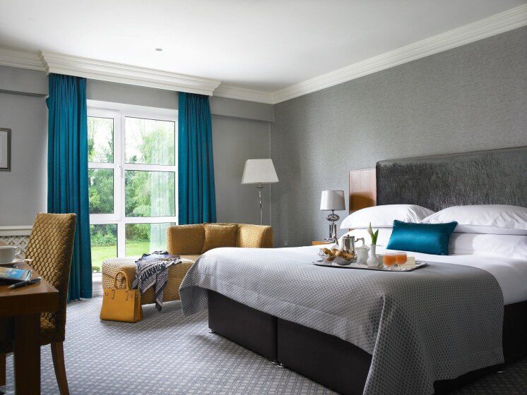 A cozy hotel room with a large bed, gray headboard, teal pillow, and bedspread. Natural light from a window with teal curtains illuminates the wicker chairs, table, and lamp. A tray with breakfast items is on the bed, creating a welcoming atmosphere.