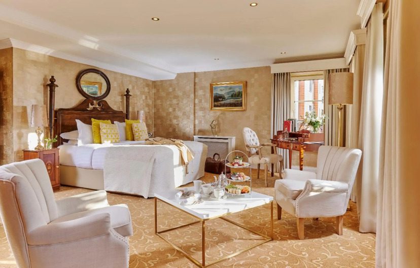 Elegant hotel room with a king-sized bed, plush armchairs, and a coffee table set with tea and pastries. Sunlit space features warm tones, luxurious decor.