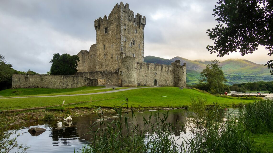 A medieval stone castle stands prominently on lush green grass, with a serene pond in the foreground and misty mountains under cloudy skies in the background.