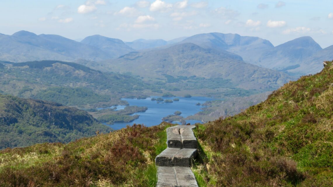 A wooden path leads through grassy hills towards a serene lake surrounded by majestic blue mountains under a clear sky, evoking tranquility.