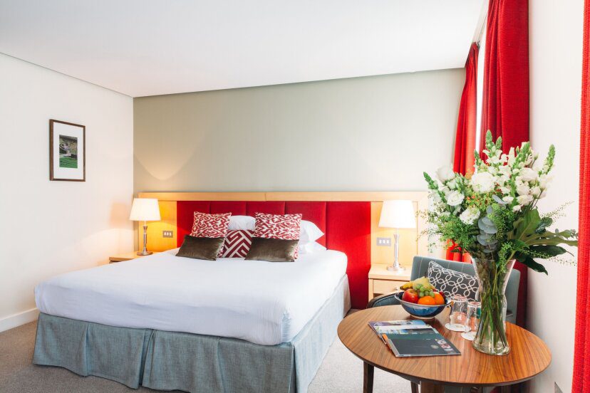A cozy hotel room with a large bed, vibrant red accents, and decorative pillows. Two lamps lit on bedside tables, and a table with fresh flowers and fruit adds elegance.