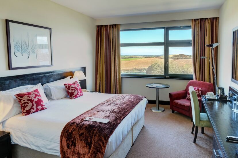 Hotel room with a large bed, adorned with white sheets and red pillows. A window with scenic countryside views, a table, and chairs create a cozy atmosphere.