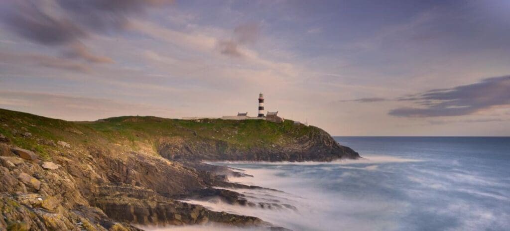 A scenic view of a rocky coastline with a tall, striped lighthouse on a grassy cliff. Gentle waves crash below under a partly cloudy sky at dusk.
