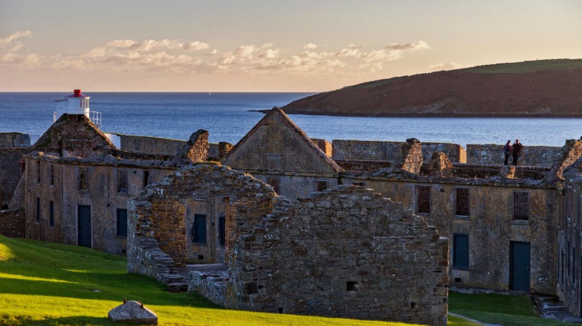Ancient stone ruins overlook a calm sea under a cloudy sky. A distant lighthouse sits on the left, while grassy hills and two figures appear on the right.