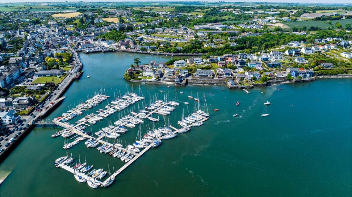 Aerial view of a vibrant marina with numerous white sailboats docked in a calm harbor. Surrounding area features lush greenery and a quaint town.
