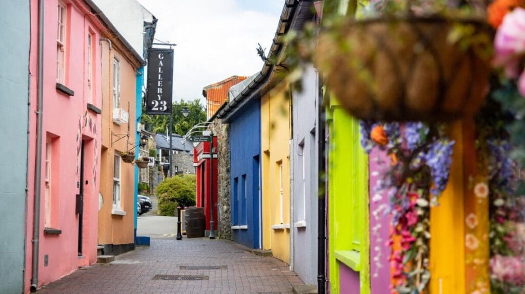 A colorful narrow street lined with vibrant houses in shades of pink, orange, blue, and yellow. A hanging basket of flowers adds charm. The scene is lively and inviting.